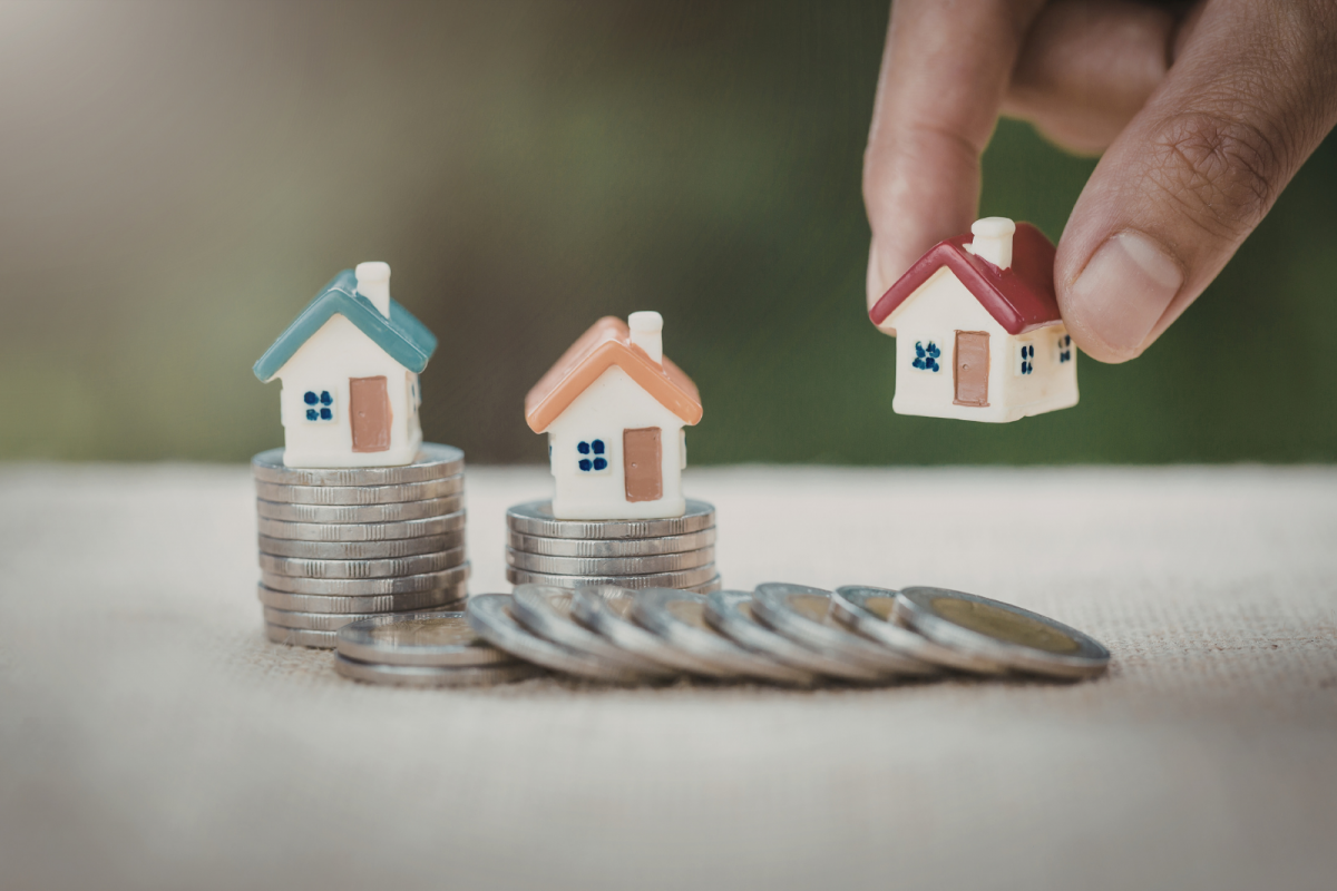 Stacked coins with miniature houses and a hand placing another property model, representing global real estate investment trends and strategic positioning for high-net-worth investors in the 2026 macro outlook.