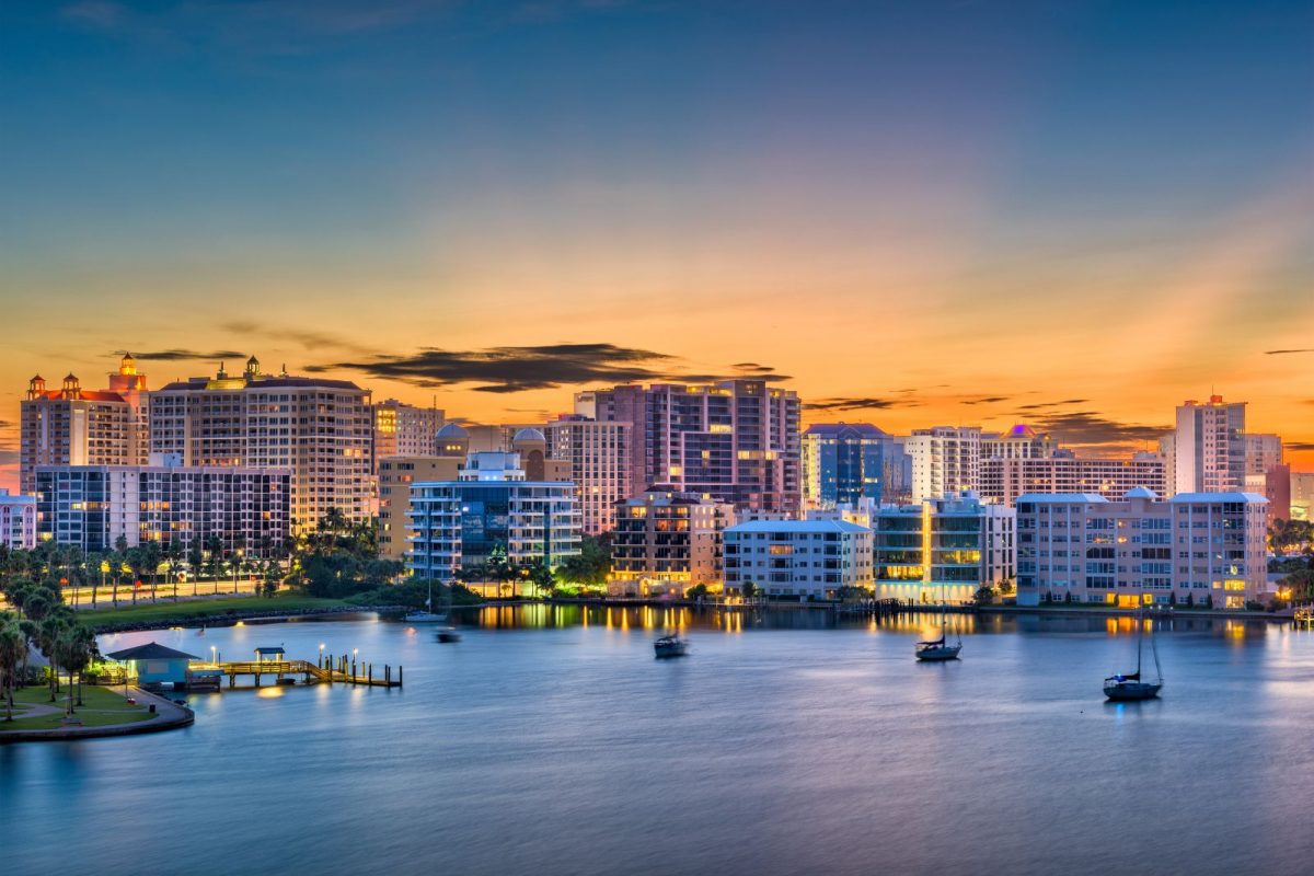 A panoramic waterfront view of the Sarasota, Florida, skyline at sunset, featuring mid-rise luxury buildings reflecting on the calm waters of the bay.
