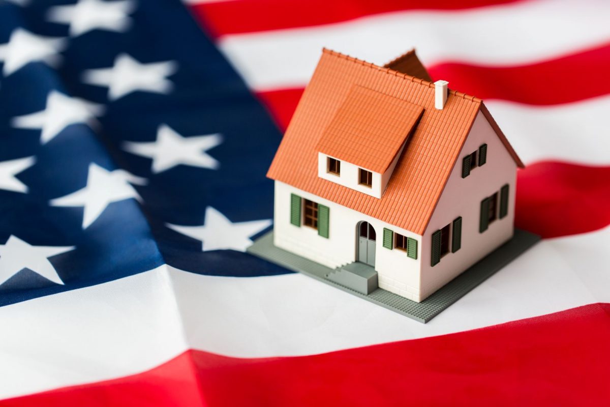 Close-up of a house model against the American flag, symbolizing high-value U.S. real estate holdings and the challenge of accessing equity within the American banking system.